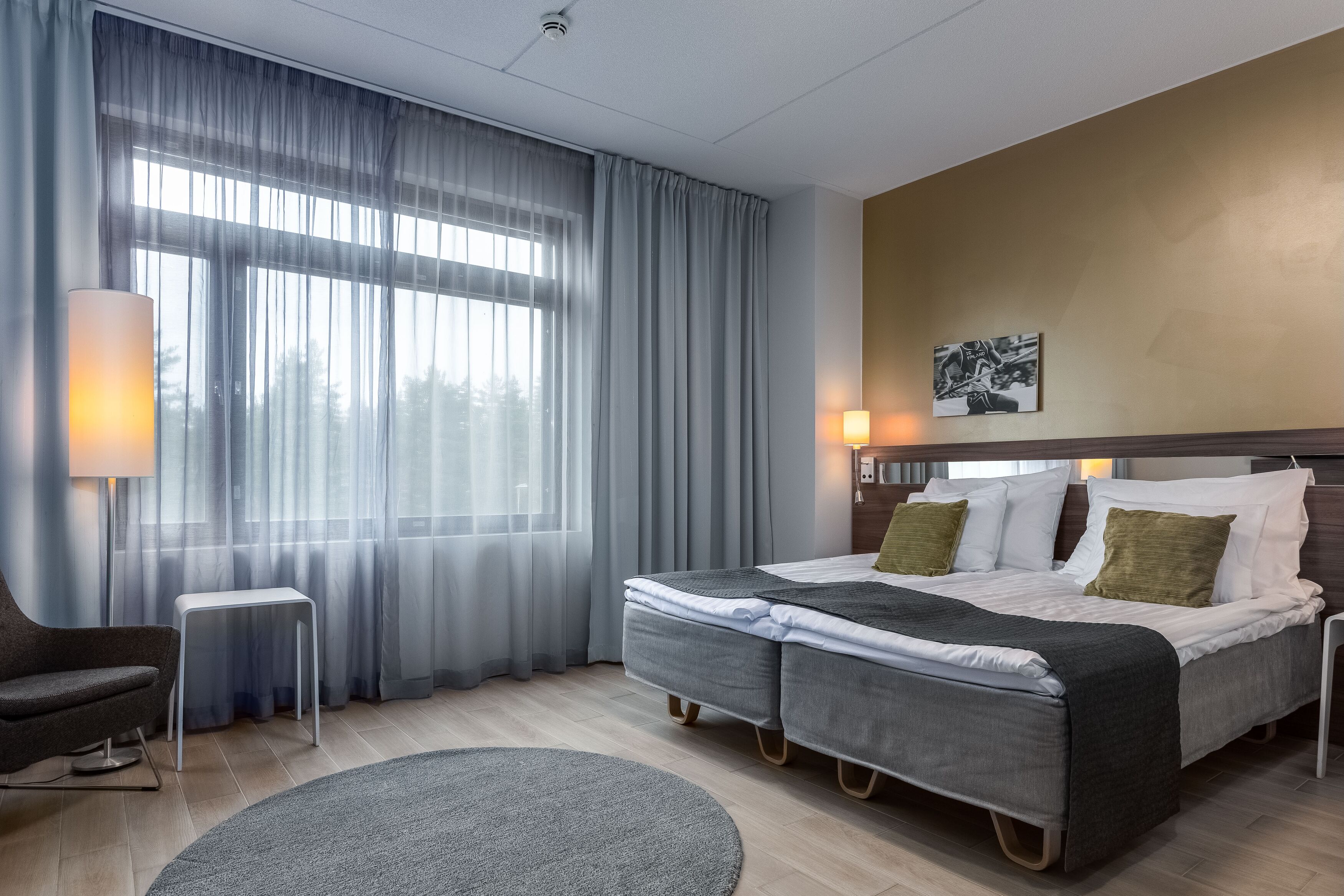 Ett modernt hotellrum med en stor säng, ett sidobord, en stol och utsikt över omgivningarna genom tunna gardiner.