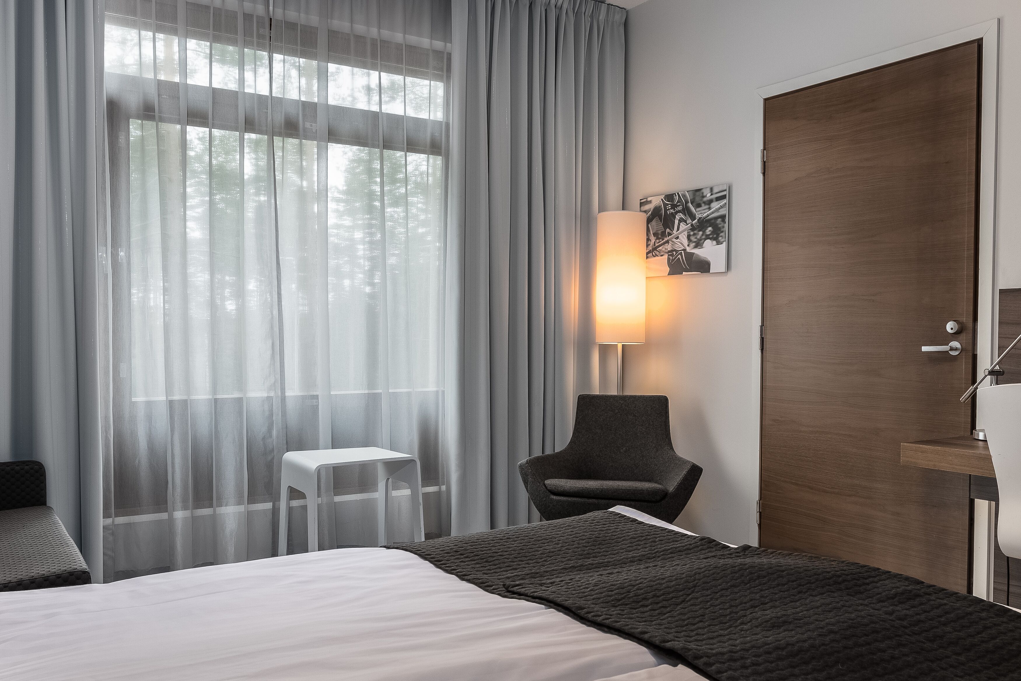A hotel room with a bed, a chair, a small table, a lamp, and a door.
