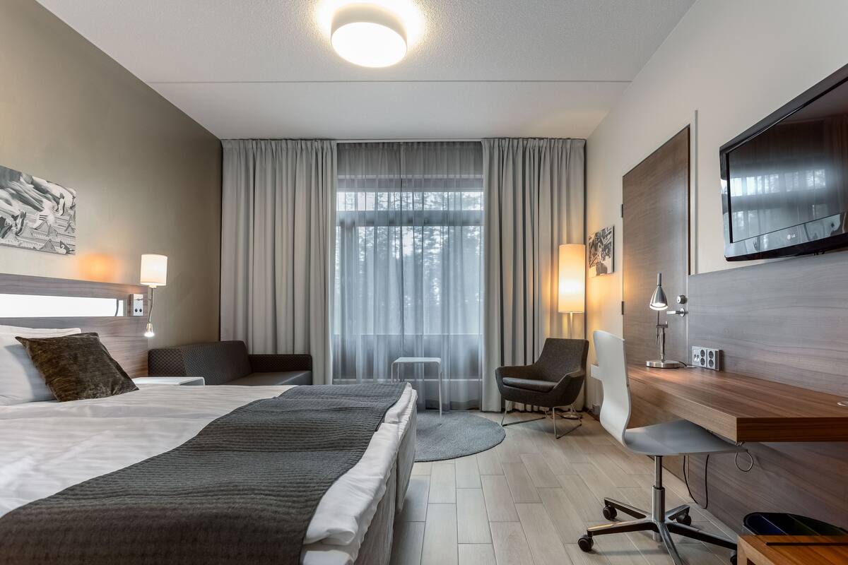 Ein Hotelzimmer mit einem großen Bett, einem Schreibtisch mit Stuhl, einem an der Wand befestigten Fernseher und einem Fenster mit Vorhängen.