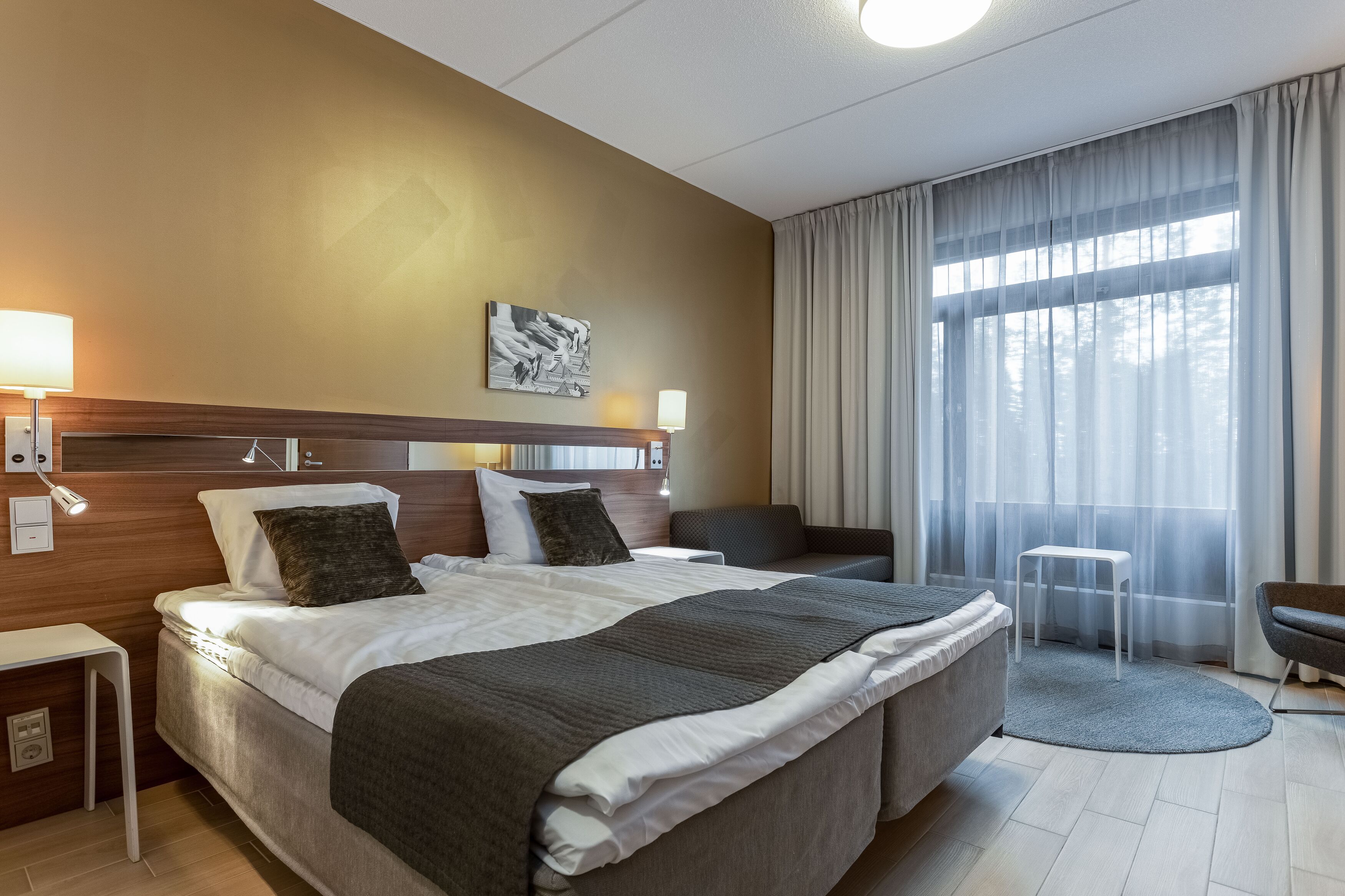 Quarto de hotel com cama grande, mesas de cabeceira, escrivaninha, cadeira e vista para o exterior através de cortinas translúcidas.