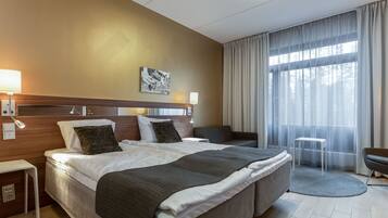 Quarto de hotel com cama grande, mesas de cabeceira, escrivaninha, cadeira e vista para o exterior através de cortinas translúcidas.