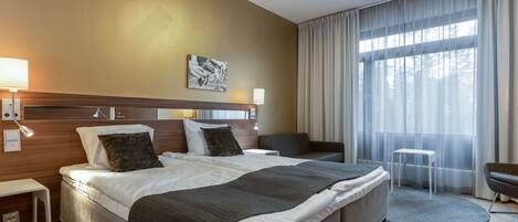 Habitación de hotel con una cama grande, mesitas de noche, un escritorio, una silla y vistas al exterior a través de cortinas translúcidas.