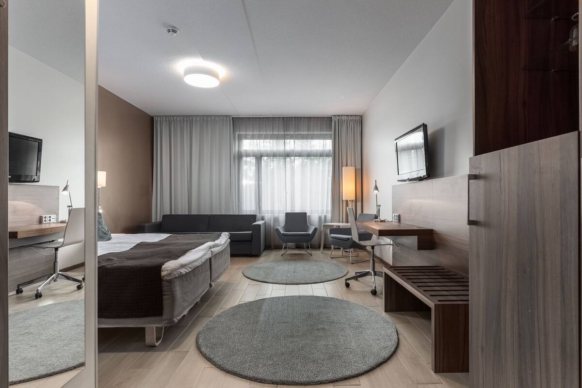 Ein modernes Hotelzimmer mit einem Bett, zwei Sesseln, einem Schreibtisch, einem Fernseher und einem großen Fenster mit Vorhängen.