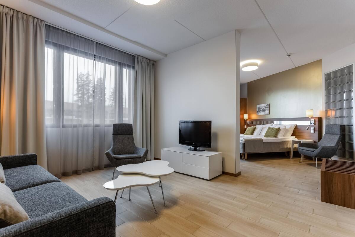 Ein modernes Wohnzimmer mit einem Flachbildfernseher, grauen Sofas, einem weißen Couchtisch und einem großen Fenster mit Vorhängen.