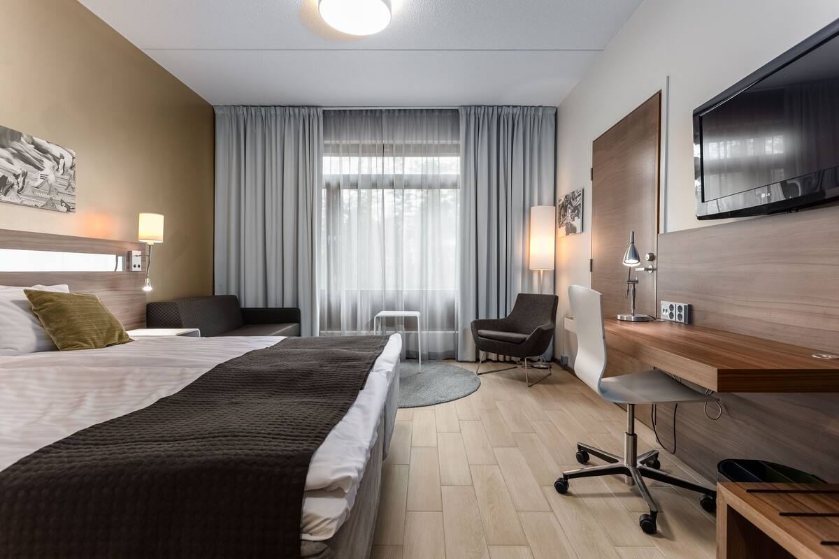 Ein Hotelzimmer mit einem großen Bett, einem Schreibtisch mit Stuhl, einem Fernseher und einem Fenster mit Vorhängen.