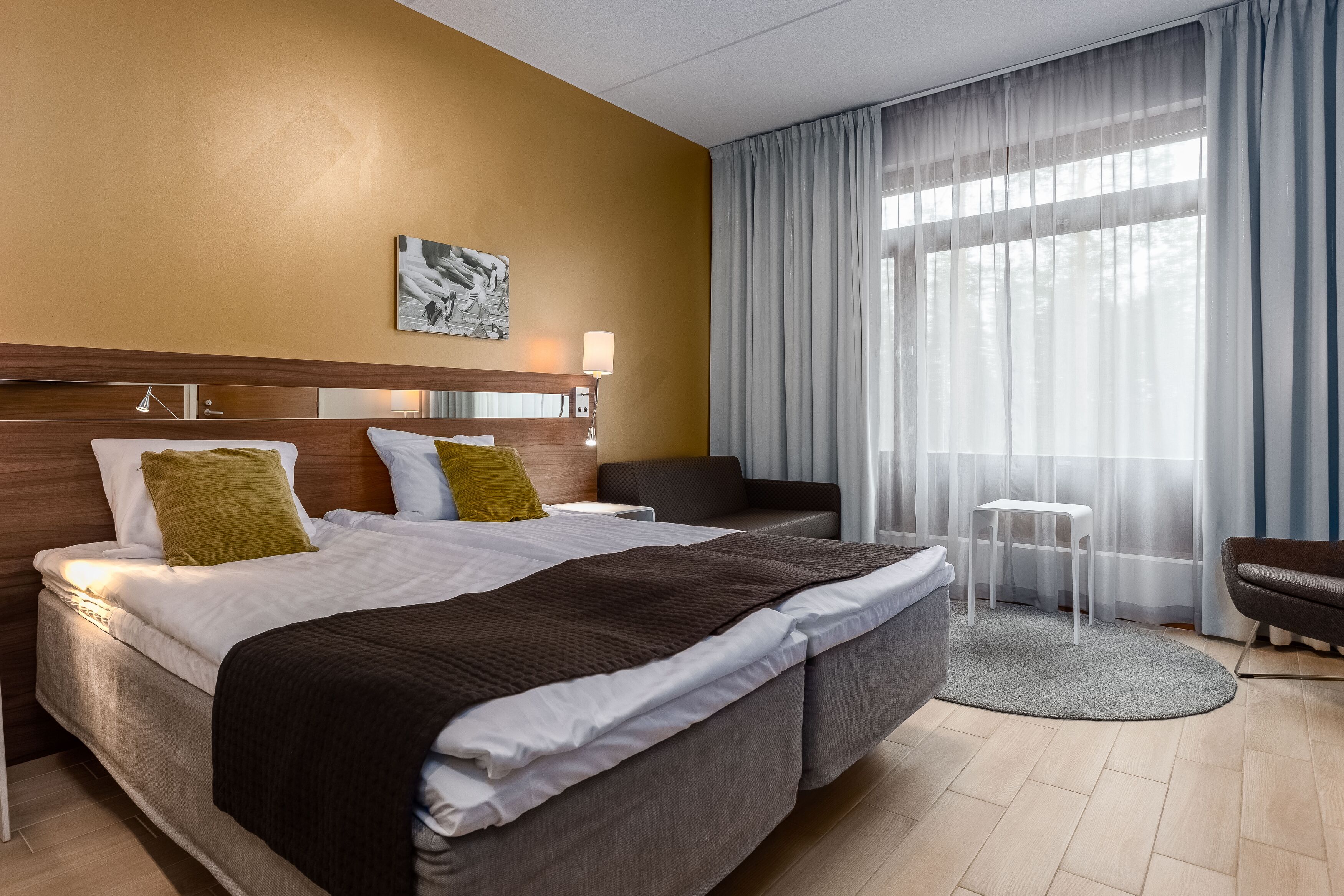 A hotel room with a large bed, a bedside table, a chair, a small table, and a window with sheer curtains.