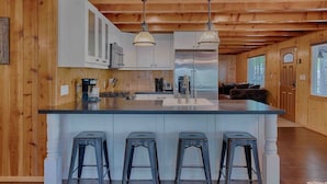 A kitchen with wooden walls, a central island with stools, pendant lights, and a view to a living area. - Super Cute Cabin in the Pines (Lakeside)