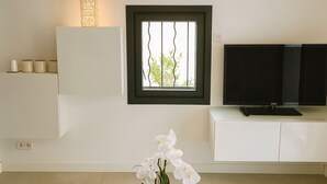 Una sala de estar moderna con un televisor sobre un mueble blanco, una ventana con vistas a follaje, y un florero con flores blancas sobre una mesa de vidrio.
