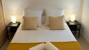 Una cama bien tendida con sábanas blancas y dos almohadas beige, flanqueada por dos mesitas de noche con lámparas, en una habitación sencilla con cabecera de madera.