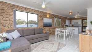 A modern living room with a grey sofa, a dining area with white chairs, and a kitchen with white cabinets.