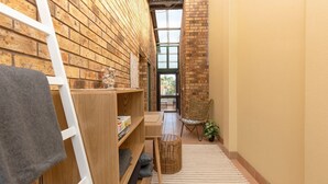 A narrow hallway with brick walls, a wooden ladder, a wicker chair, and a small table with a plant.