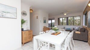 A spacious dining area with a long white table and chairs, a wooden cabinet, and a view of the outdoors through large windows.