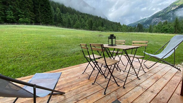 Una terraza de madera con una mesa y sillas, con vistas a un campo cubierto de hierba y montañas boscosas.