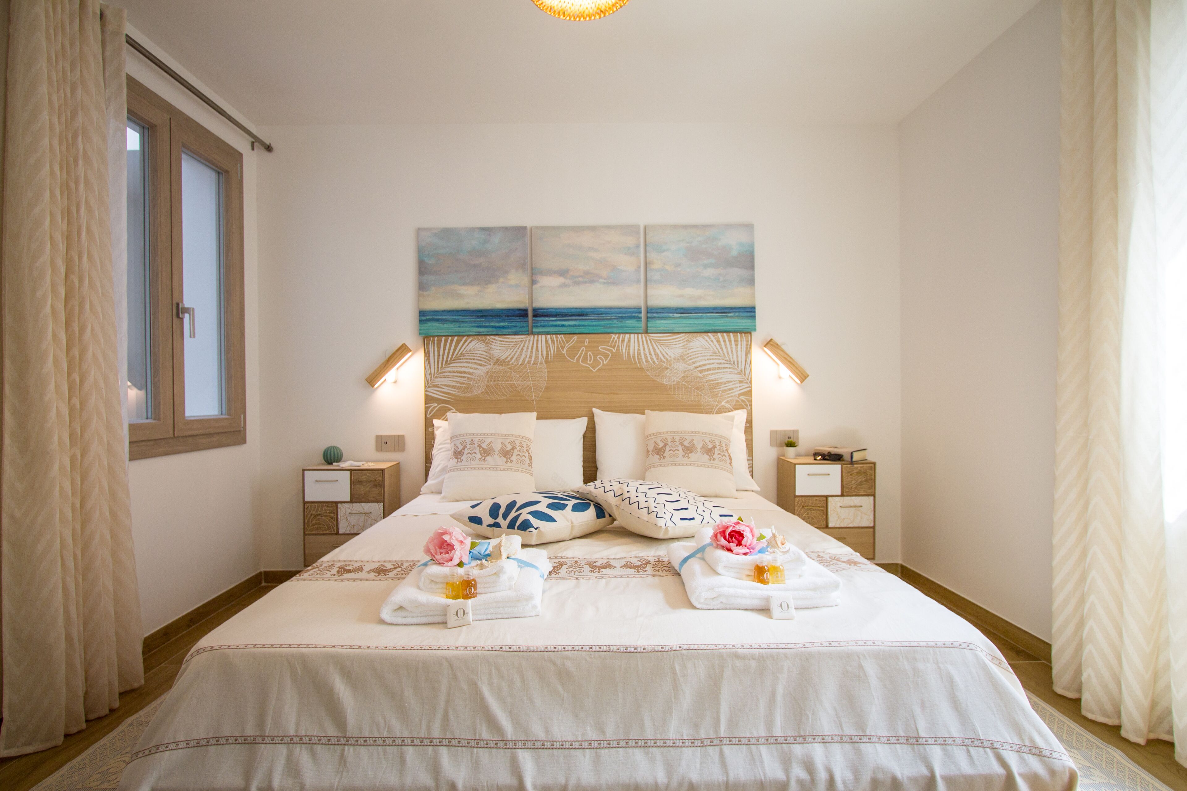 A neatly made bed with a coastal artwork above it, bedside tables with lamps, and a window with curtains.