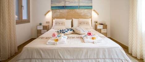 A neatly made bed with a coastal artwork above it, bedside tables with lamps, and a window with curtains.
