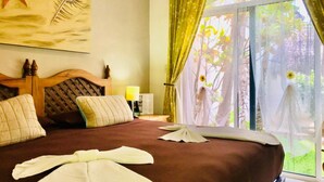 A hotel room with a large bed, a wooden headboard, a nightstand with a lamp, and a view of greenery through the window. - Double Private Villa Playacar - Pool - Walk to Beach & 5th Avenue - Sleeps 22 (Playa del Carmen)