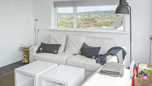 Een moderne woonkamer met een witte bank, twee witte bijzettafeltjes en een raam met uitzicht op een landelijk landschap.