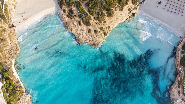 An aerial view of a coastal area with a sandy beach, clear turquoise waters, and a rocky outcrop with dense greenery. - Cala Domingos Apartments (Manacor)