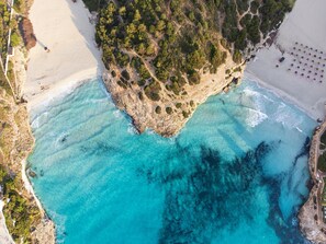 Vista aérea de una zona costera con una playa arenosa, aguas turquesas cristalinas y un promontorio rocoso con vegetación densa.