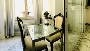 A dining area with a glass table and chairs, a statue, a window with curtains, and a shower area with a glass door.