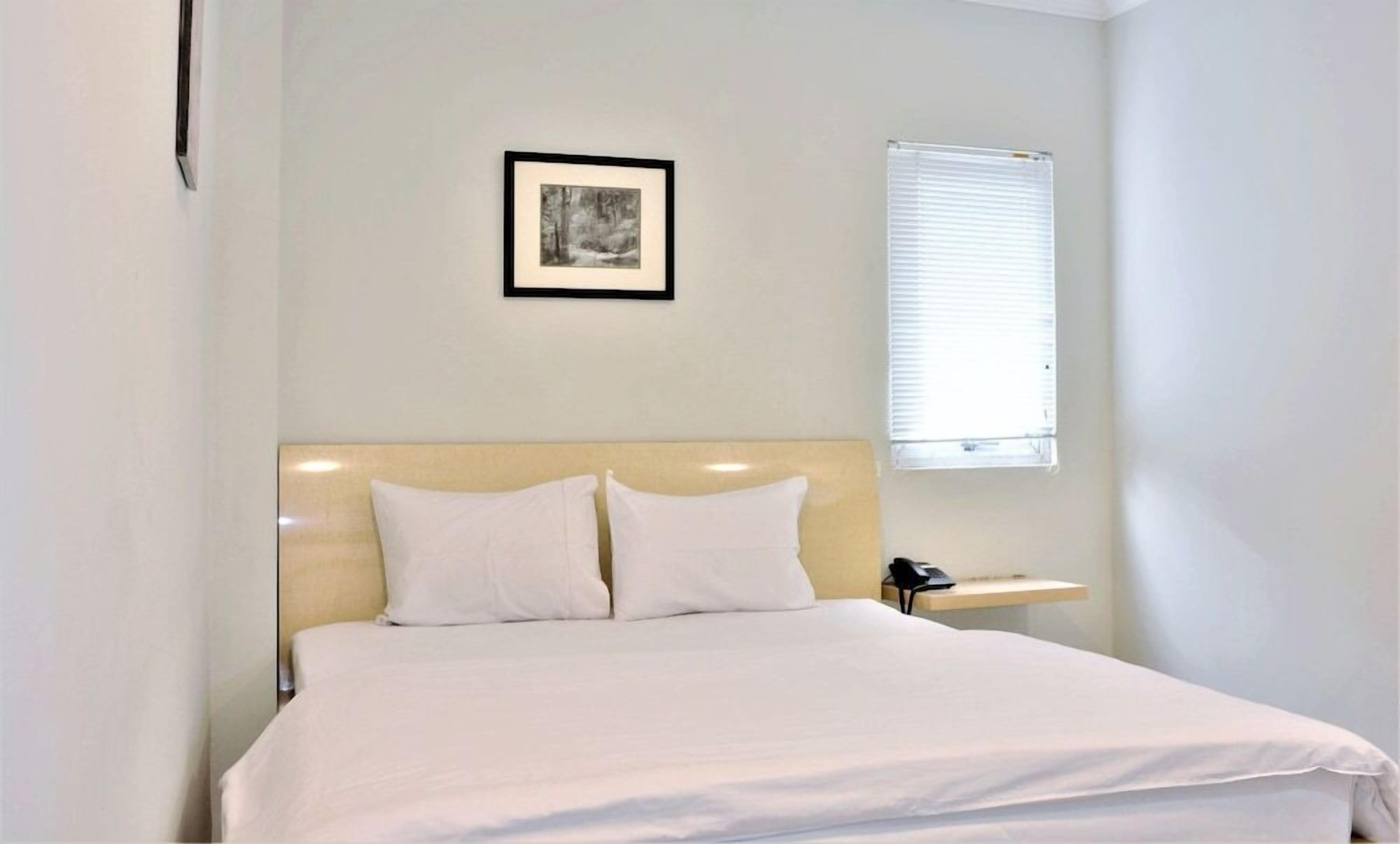 Una cama bien tendida con sábanas blancas en una habitación con pared de color claro y un cuadro enmarcado encima.