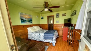 Une pièce de taille modeste comprenant un lit, une tête de lit en bois, un ventilateur de plafond, un tableau, une horloge et un petit meuble.