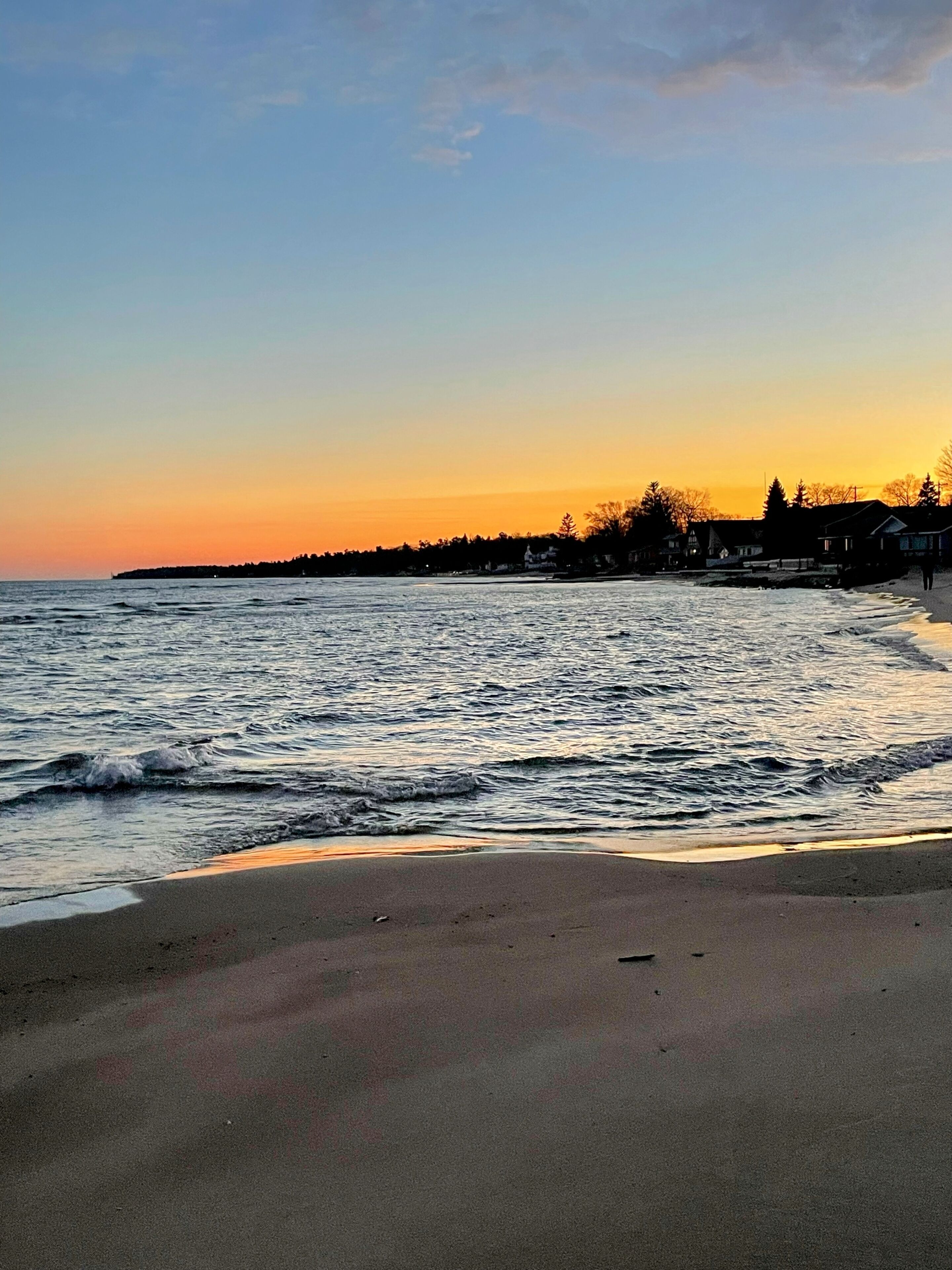 A beach with waves at sunset, a distant shoreline with houses, and a clear sky.