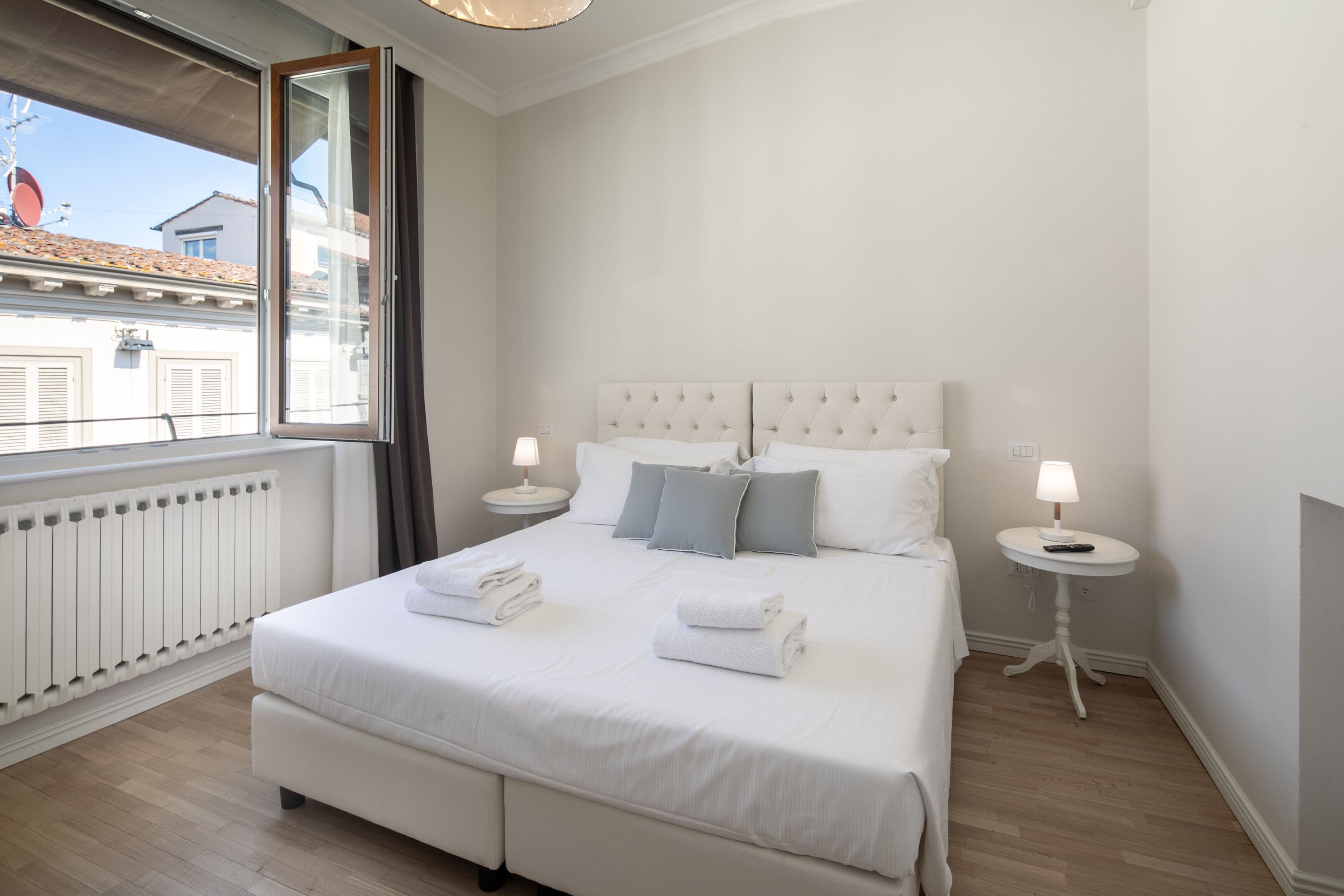 A neatly made bed with white linens and folded towels, a window with a view of buildings, and two bedside tables with lamps.