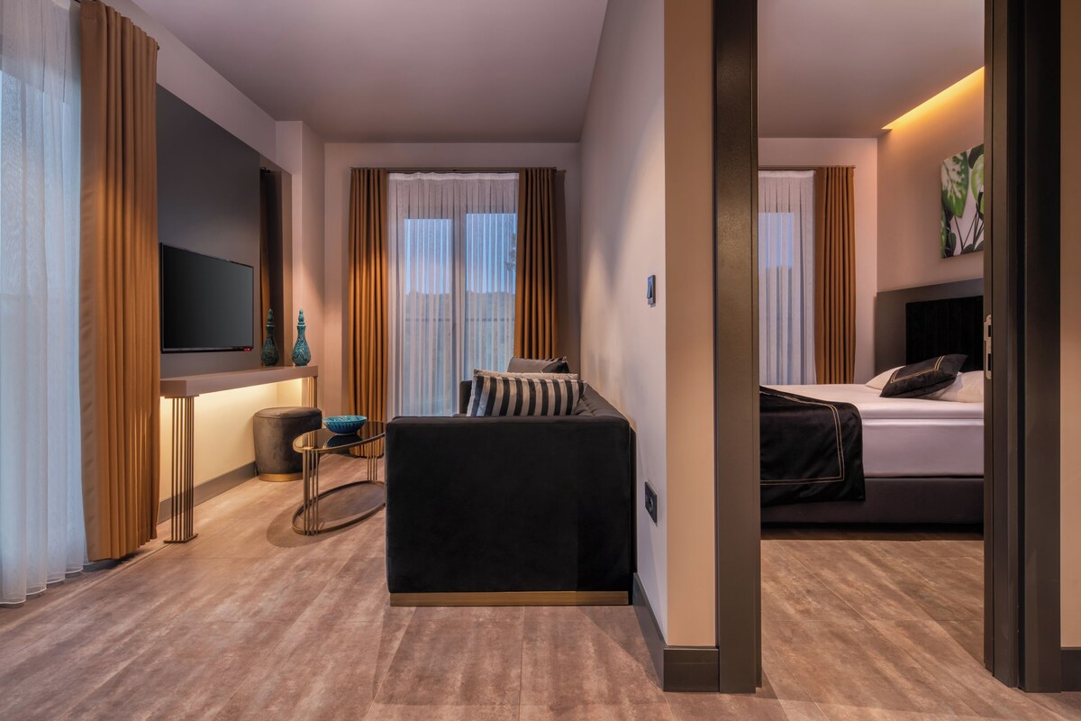 A modern hotel room with a large bed, a sofa, a TV mounted on the wall, and a view of the city through large windows.