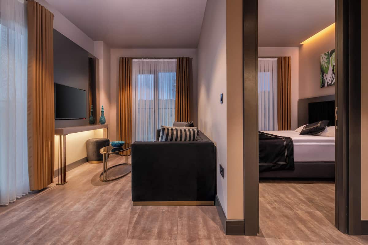Una habitación de hotel moderna con una cama grande, un sofá, un televisor montado en la pared y vistas a la ciudad a través de amplios ventanales.