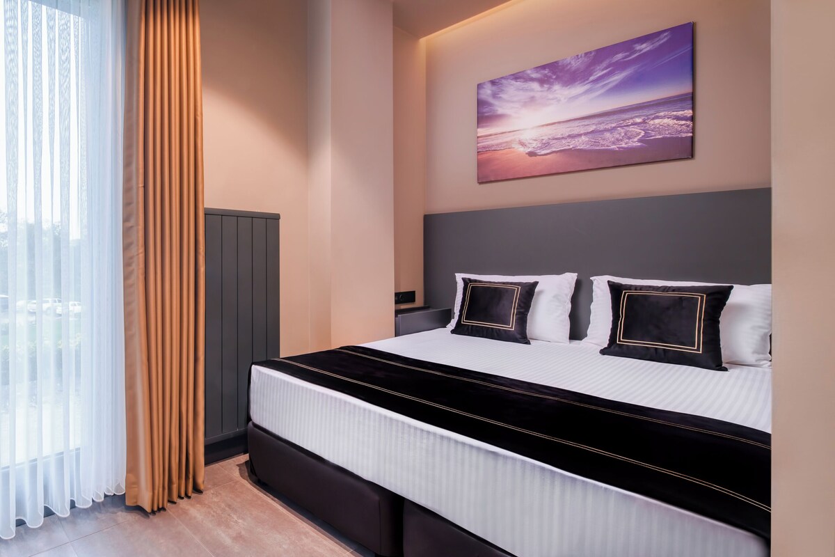 A modern hotel room with a large bed, a view of the outdoors through sheer curtains, and a painting on the wall.