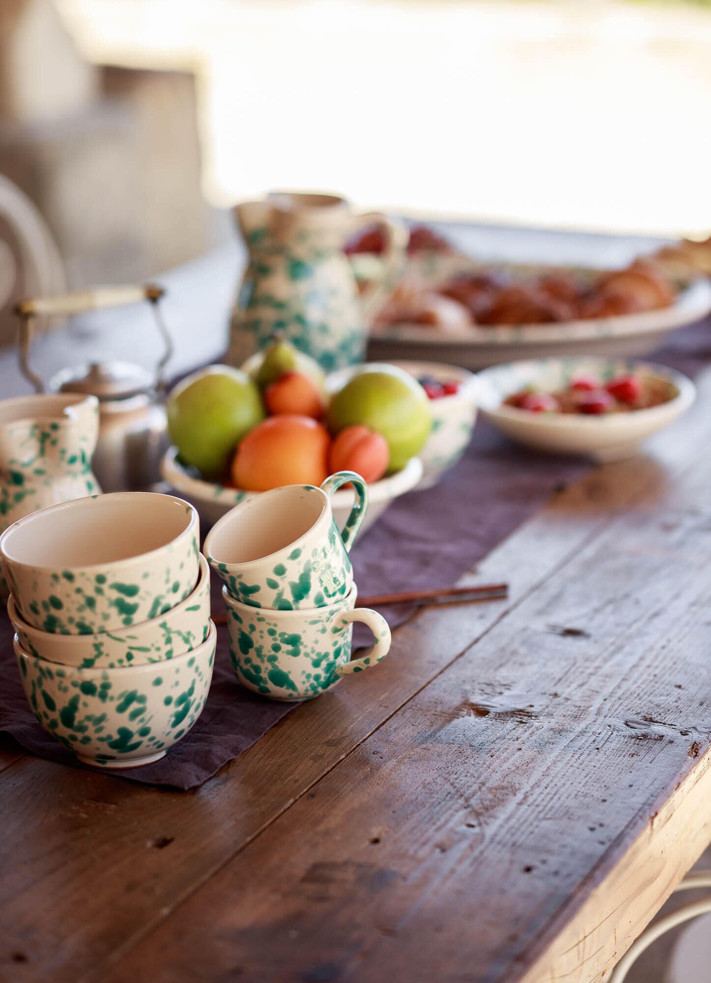 Une table en bois avec des tasses en céramique et un pichet, un saladier rempli de fruits et une assiette de pâtisseries.