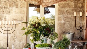 Un área de comedor al aire libre con una mesa de madera rústica, sillas de metal blanco, una lámpara colgante tejida, un centro de mesa floral y un fondo de pared de piedra.