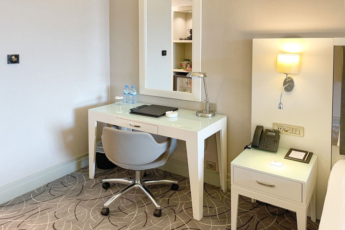 Quarto de hotel com escrivaninha branca, cadeira e mesa de cabeceira, equipado com luminária, telefone e garrafas de água.