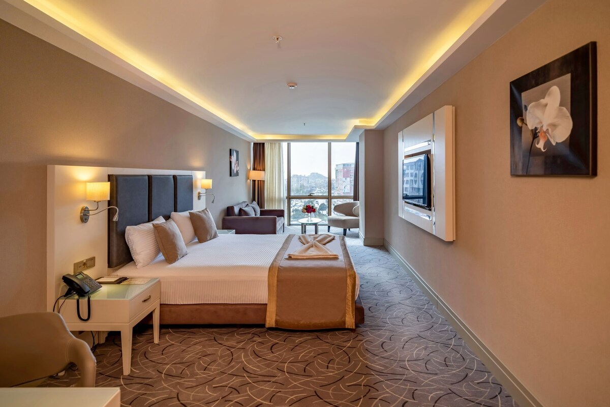 Une chambre d’hôtel avec un grand lit, des lampes de chevet, un bureau avec un téléphone, une chaise, une petite table avec un vase et une vue sur la ville grâce à de grandes fenêtres.