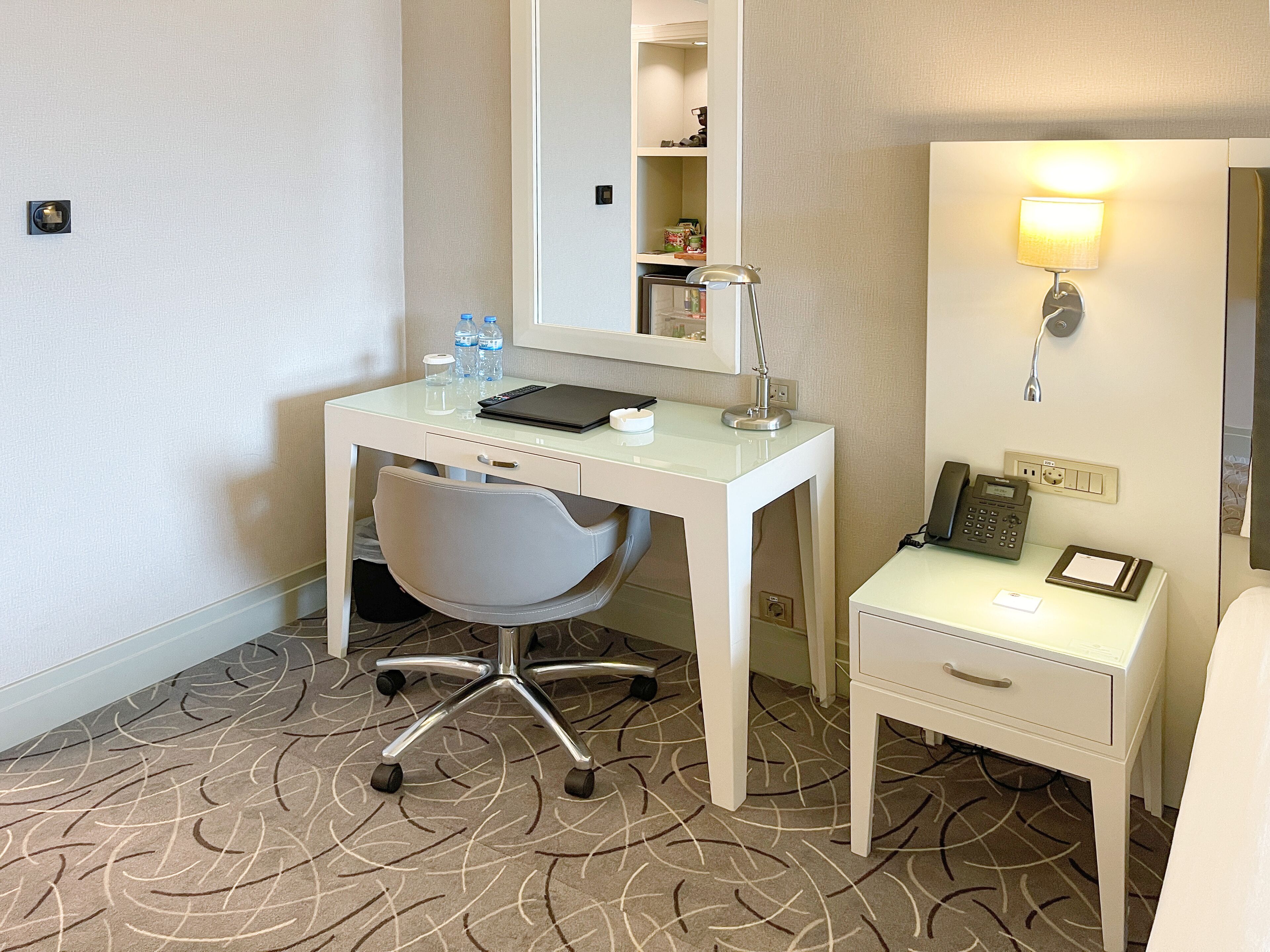Een hotelkamer met een wit bureau, een stoel, een klein nachtkastje met een lamp en een spiegel.