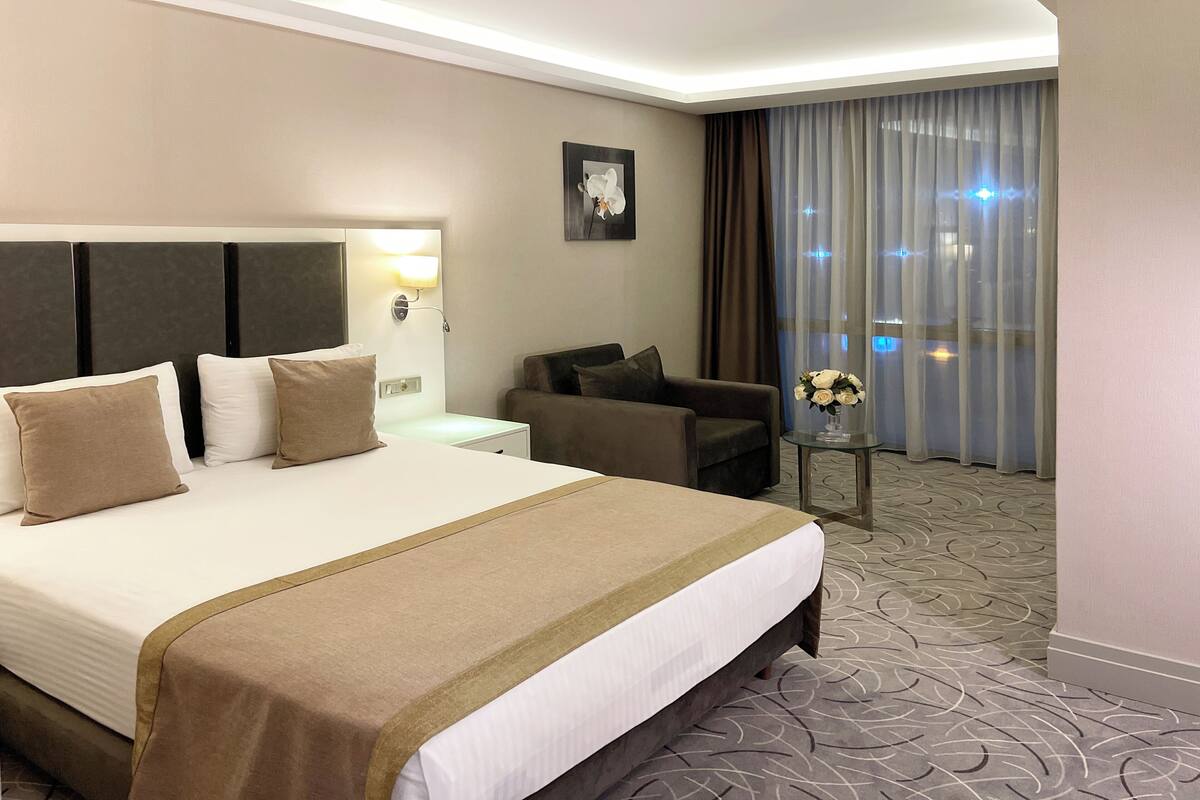 A hotel room with a large bed, a sofa, a small table with a vase, and a window with curtains.