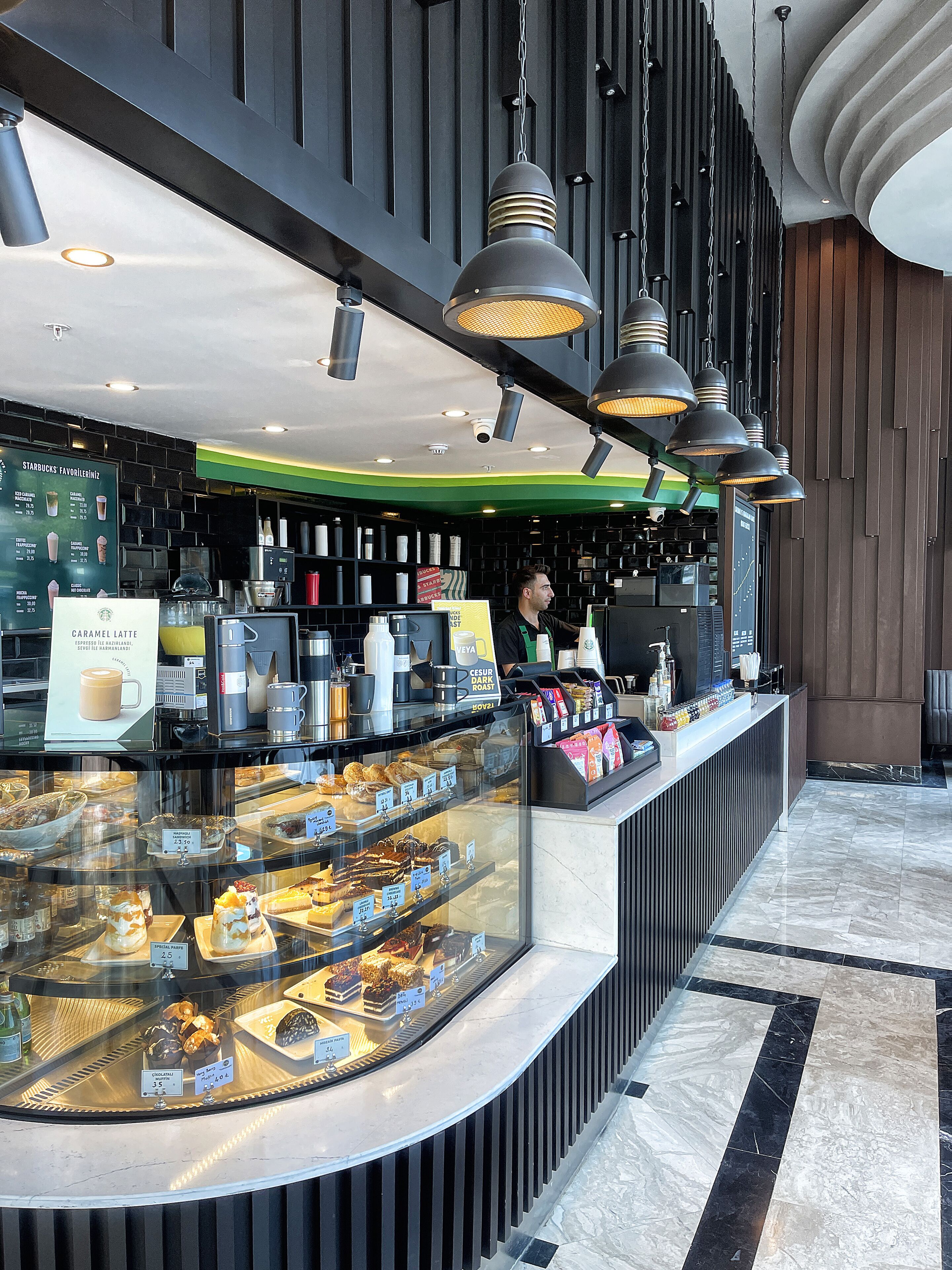 A modern coffee shop interior with a counter displaying pastries and drinks, a menu board, and a cashier.