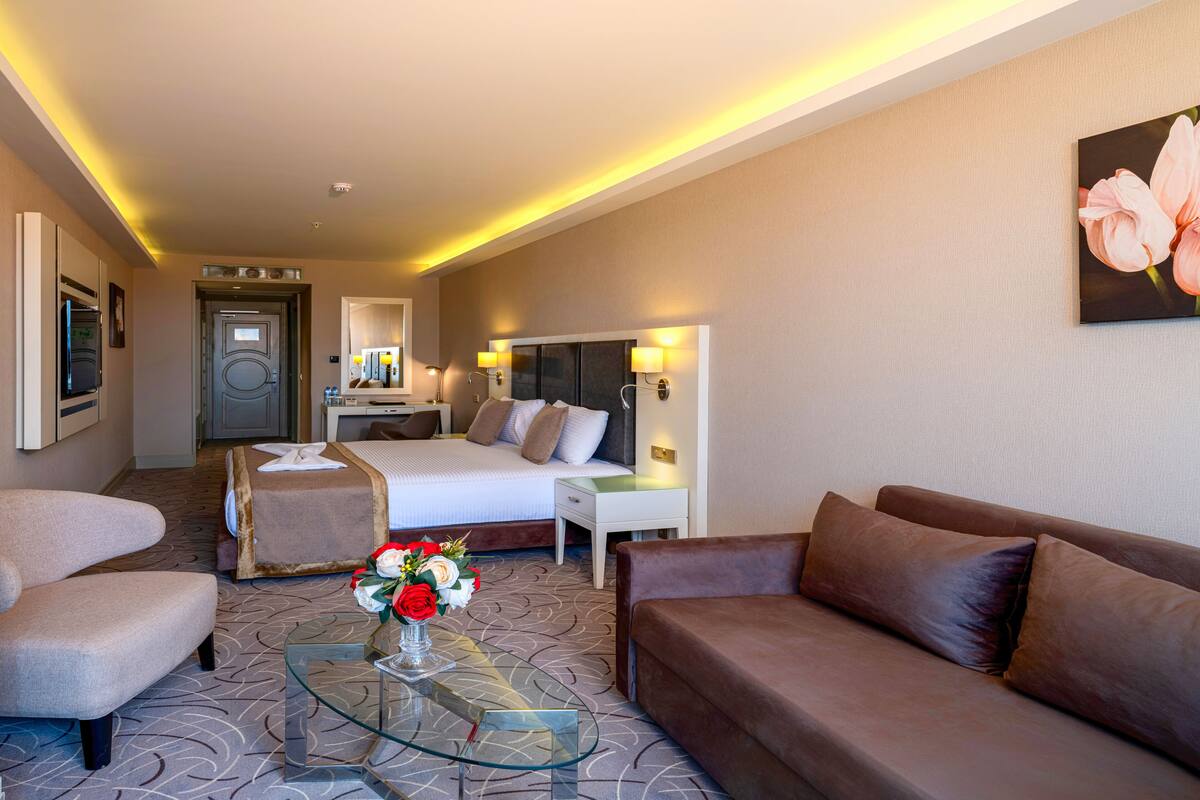 A hotel room with a bed, two armchairs, a sofa, a glass coffee table with a vase of flowers, and a painting on the wall.