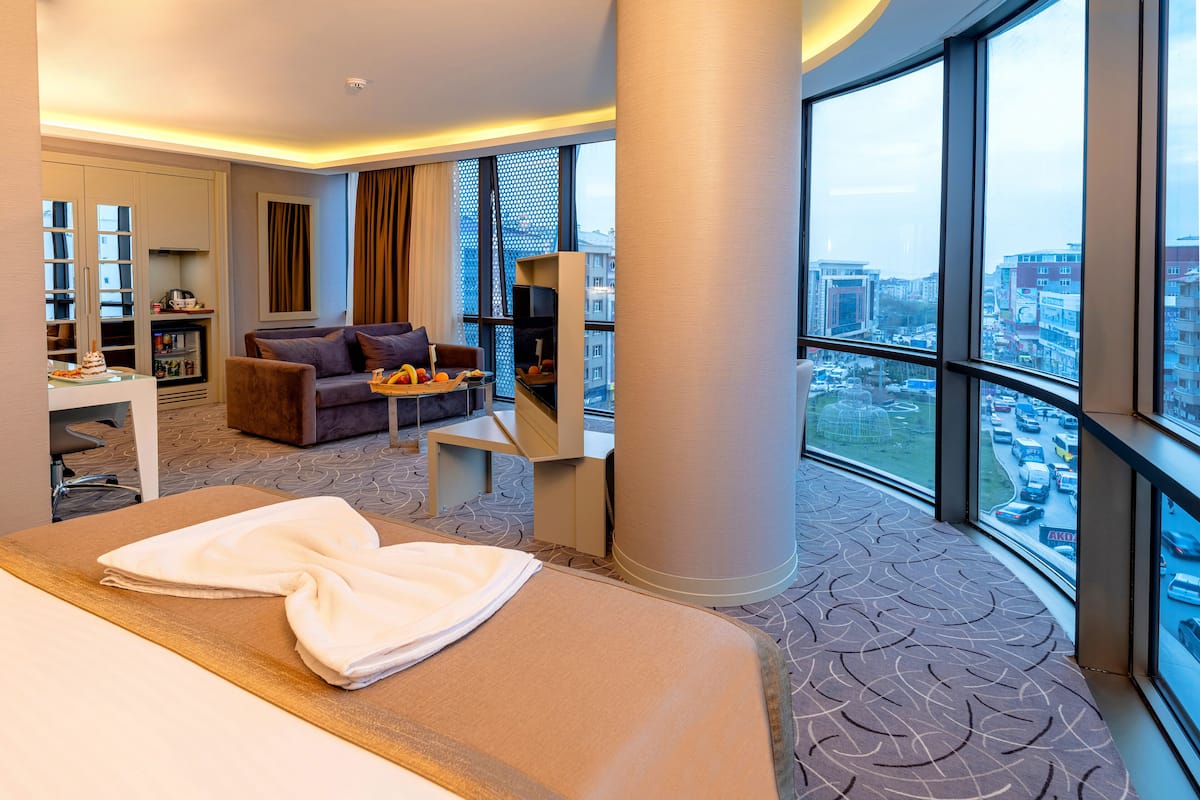 A modern hotel room with a large bed, a sofa, a small table, and a view of the city through large windows.