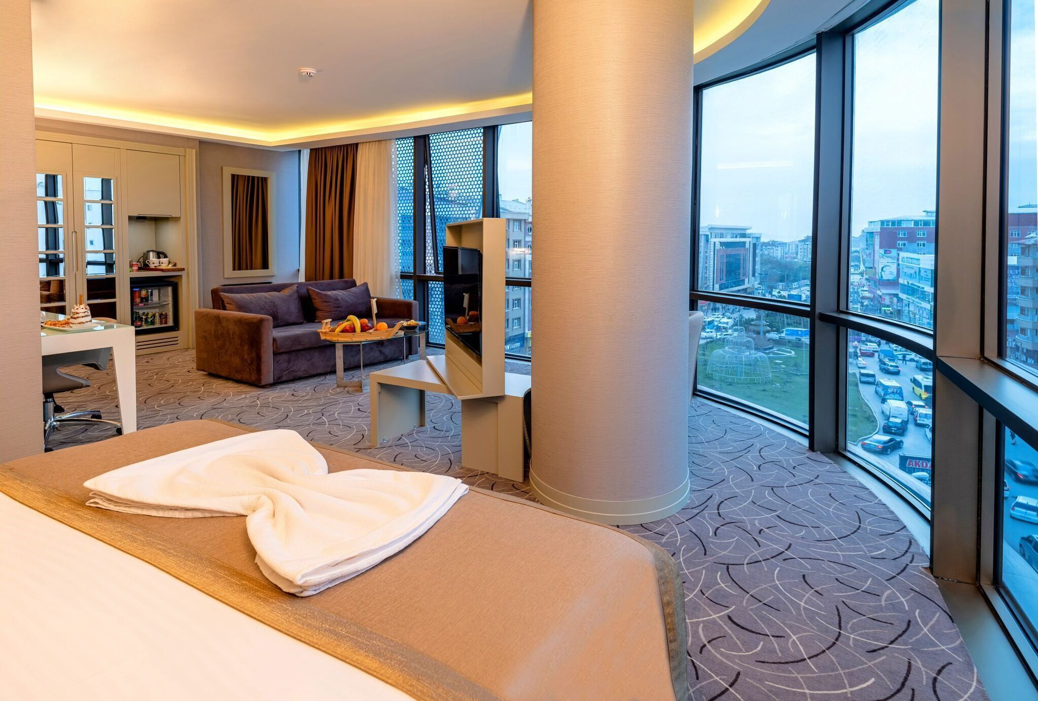 A hotel room with a bed, a sofa, a small table, a bookshelf, and a large window offering a city view.