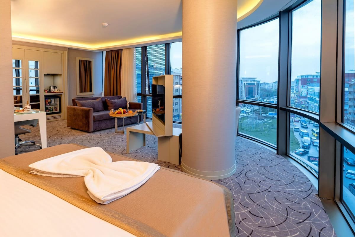 A hotel room with a bed, a sofa, a small table, a bookshelf, and a large window offering a city view.