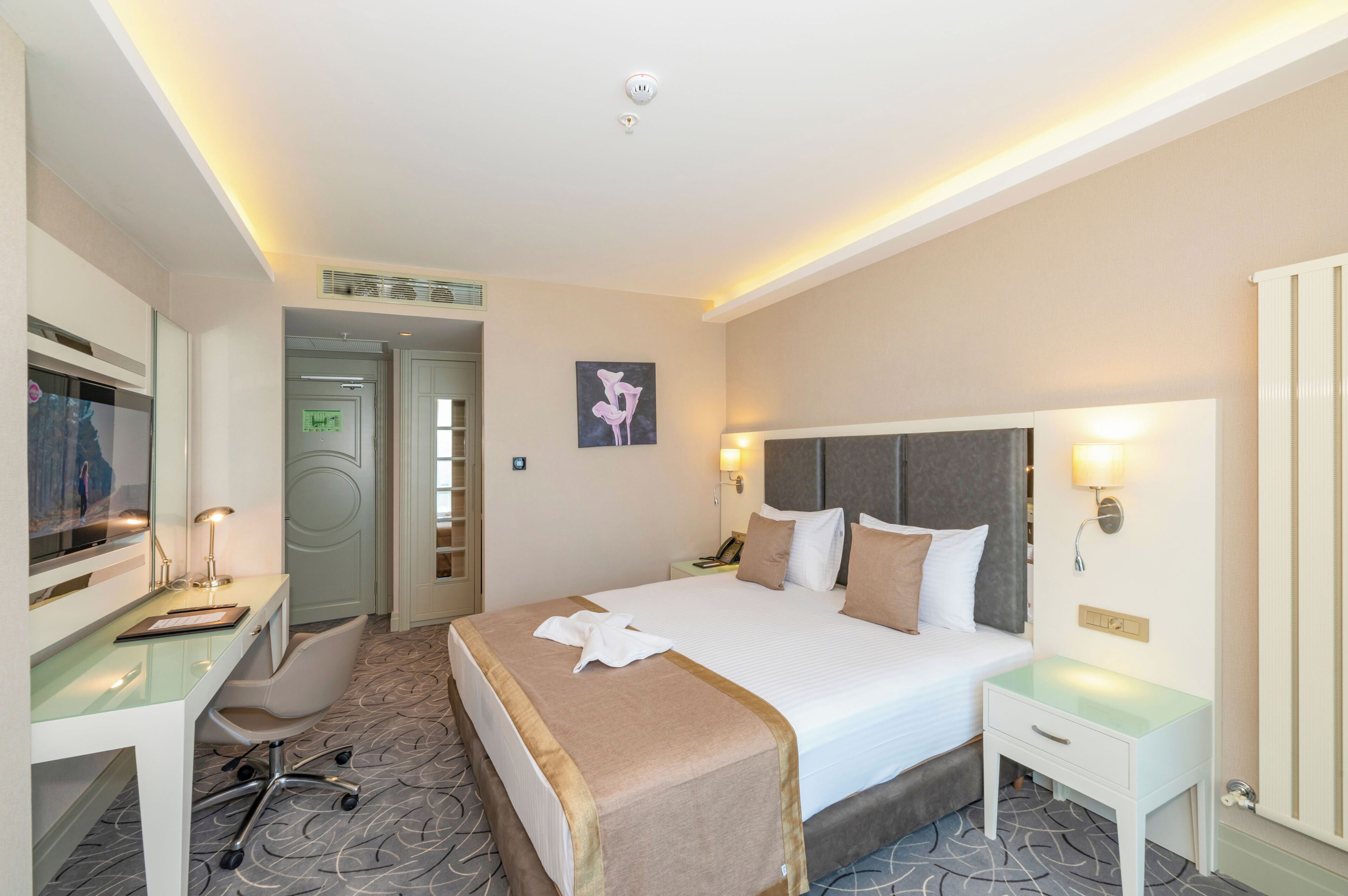 A hotel room with a large bed, a desk, a chair, a nightstand, and a wall-mounted lamp.