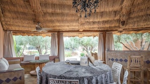 Un espace de restauration extérieur doté d’un toit de chaume, avec une table ronde, des chaises et une vue sur les arbres et la verdure.