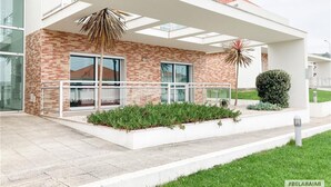 A modern building with a brick facade, large windows, and a covered outdoor area with a palm tree and a bush. - Modern, air-conditioned family apartment with pool. 10 minute walk to beach (Salir do Porto)