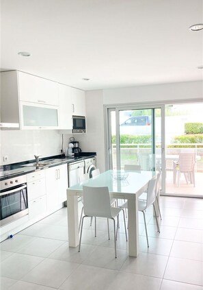 A modern kitchen with white cabinets, a glass dining table, and a view of the outdoors through a sliding glass door. - Modern, air-conditioned family apartment with pool. 10 minute walk to beach (Salir do Porto)