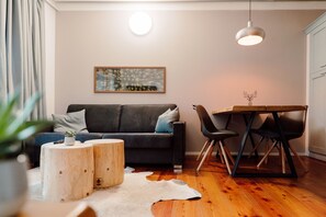 A modern living room with a dark grey sofa, wooden side tables, a dining table with chairs, a white rug, and a painting on the wall.