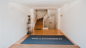 Ein Flur mit Holzboden, der zu einer Wendeltreppe führt. Eine Fußmatte mit dem Wort 'WILLKOMMEN' liegt am Eingang.