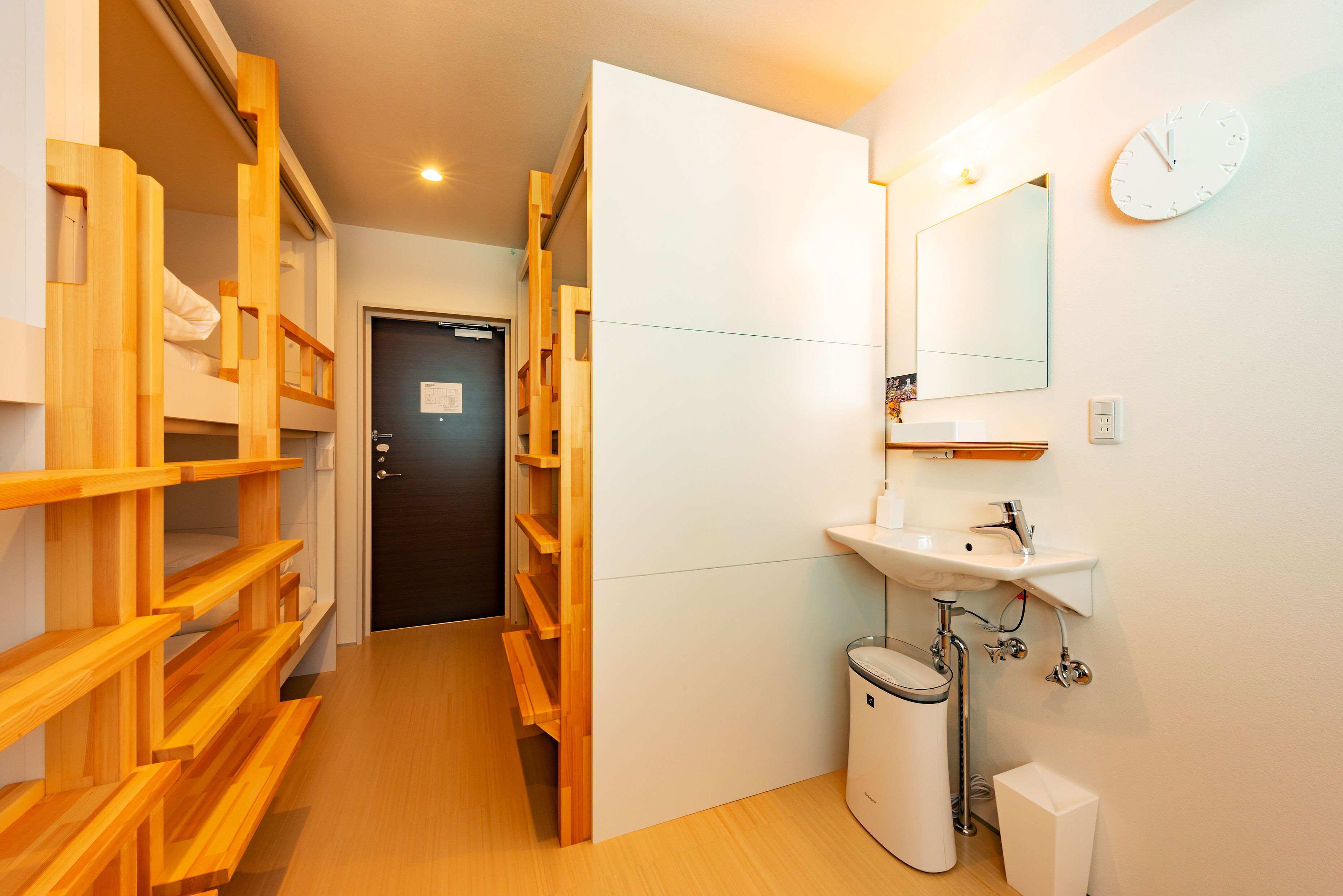 Una habitación de hotel con una cama litera de madera, un lavamanos blanco, un espejo y un reloj en la pared.