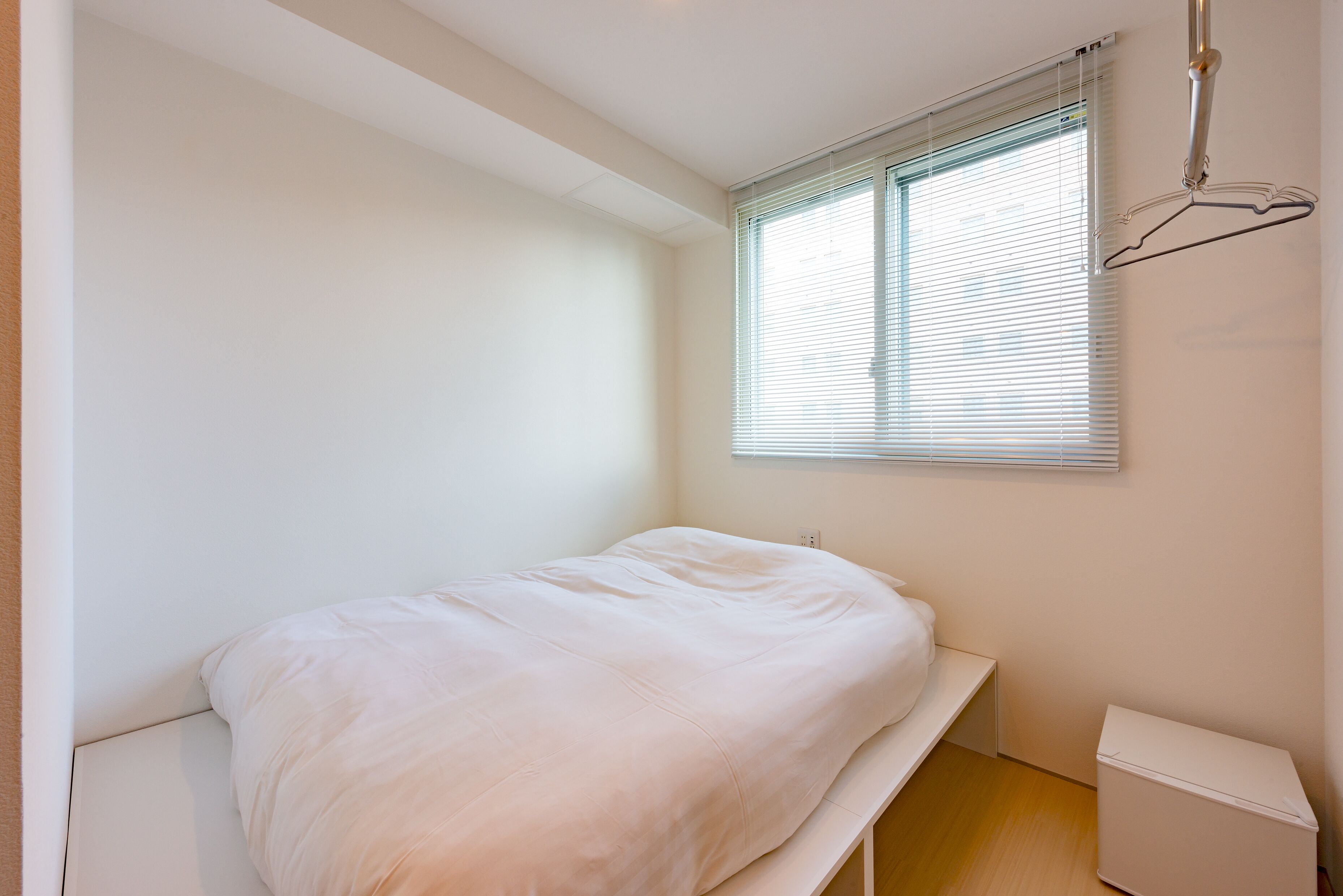 A small, neatly arranged bedroom with a single bed, a bedside table, and a window with blinds.
